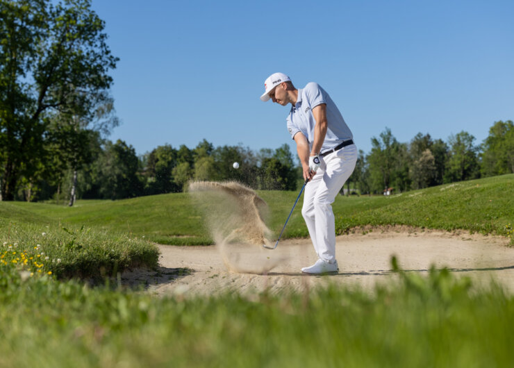 Golfer i et sandfang slår et slag, sandet flyver, grønne træer og blå himmel i baggrunden.