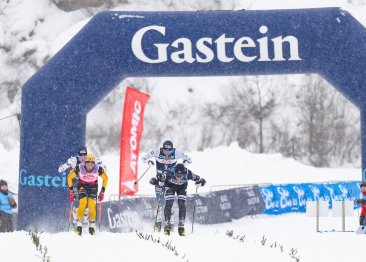©Gasteinertal Tourismus GmbH, Mangotree Photography, GasteinCriterium Langrendsløbere løber under en buegang i Gastein på en snedækket bane med tilskuere i baggrunden.