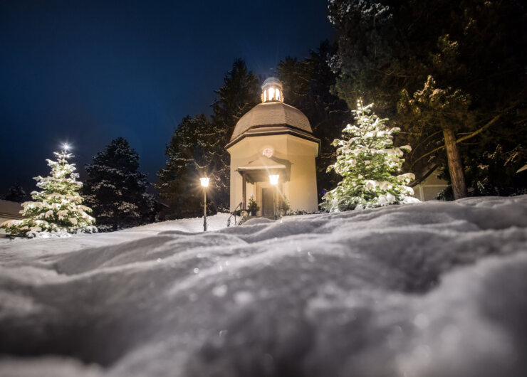 Et lille kapel lyser op om natten, omgivet af snedækkede træer og oplyste gadelamper.