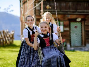 Drei Mädchen in traditionellen Kleidern schaukeln während des Bauernherbstes vor einem Holzgebäude und lächeln dabei fröhlich.