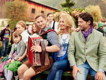 Eine Gruppe in traditioneller österreichischer Kleidung feiert den Bauernherbst im Freien, während ein Mann Akkordeon spielt.