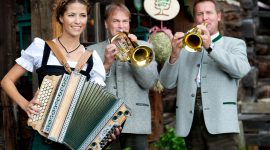 Drei Personen in traditioneller österreichischer Kleidung spielen Akkordeon und Trompeten bei einem rustikalen Bauernherbstfest.