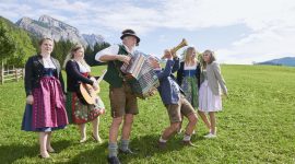 Menschen in traditioneller österreichischer Kleidung feiern den Bauernherbst mit Musik und Gesang auf einer Wiese mit den Bergen im Hintergrund.