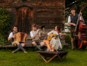 Vier Musikanten in traditioneller österreichischer Kleidung spielen Instrumente an einer Bauernherbst-Feuerstelle bei einer rustikalen Holzhütte.