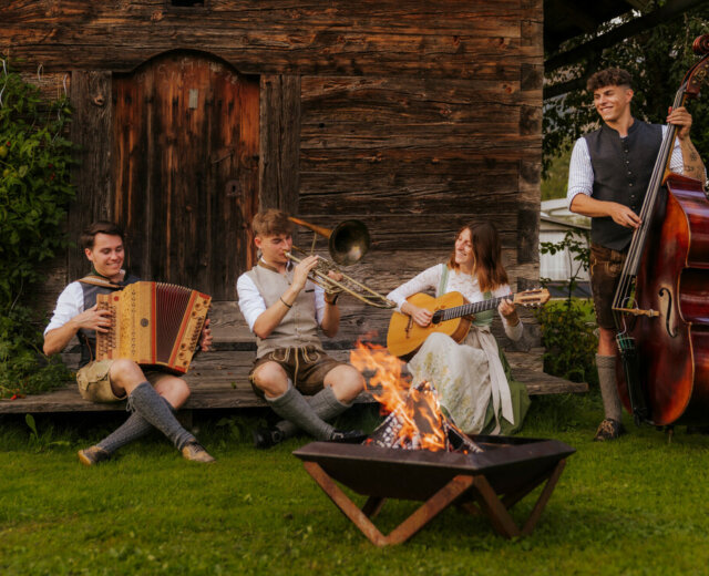 Musik im Bauernherbst Vier Musikanten in traditioneller österreichischer Kleidung spielen Instrumente an einer Bauernherbst-Feuerstelle bei einer rustikalen Holzhütte.