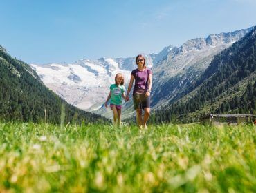 Frau und Kind halten sich an den Händen und gehen auf einer Wiese im Krimmler Tal mit schneebedeckten Gipfeln im Hintergrund.