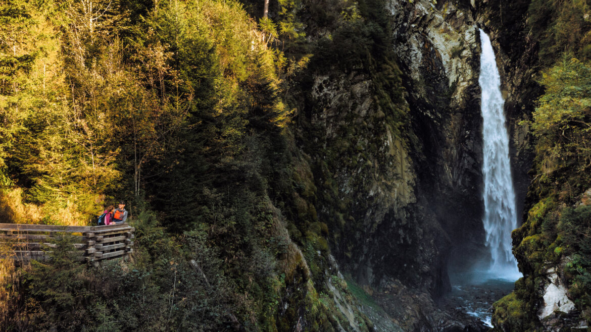 Menschen stehen auf einer Holzplattform in der Nähe eines hohen Wasserfalls in der üppig bewaldeten Schlucht von Neukirchen. (vergrößerte Ansicht)