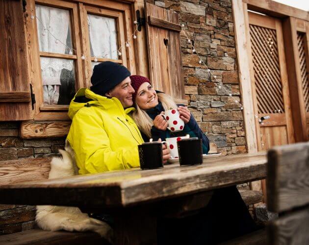 Ein lächelndes Paar in Winterkleidung genießt einen Hüttenurlaub mit einer gepunkteten Tasse vor einer rustikalen Hütte.