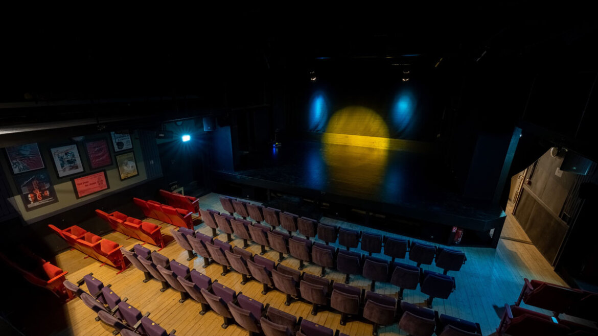 Leeres Theater in Neukirchen mit lila und roten Sitzen vor einer dunklen, von bunten Scheinwerfern beleuchteten Bühne. (vergrößerte Ansicht)