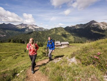 2016-08-03-SLT-Twengeralmse Zwei Wanderer gehen auf einem Bergpfad im Lungau, mit grünen Hügeln, Wildblumen und Hütten im Hintergrund.