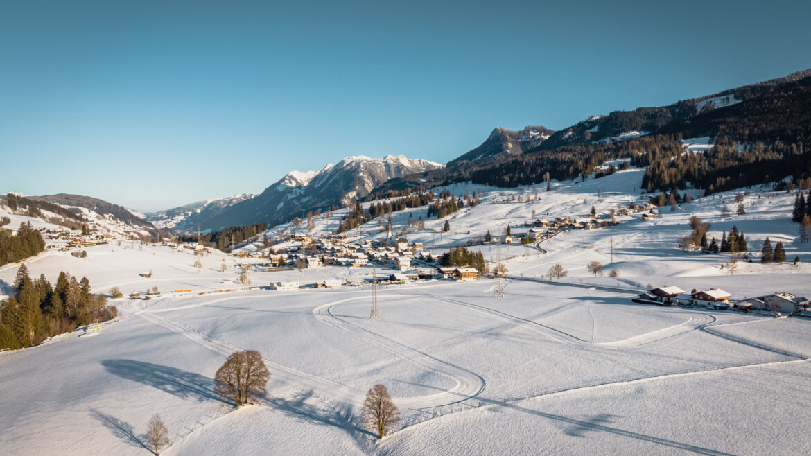 Ein verschneites Dorf Embach liegt in einem Tal mit Bergen im Hintergrund unter einem klaren blauen Himmel. (vergrößerte Ansicht)