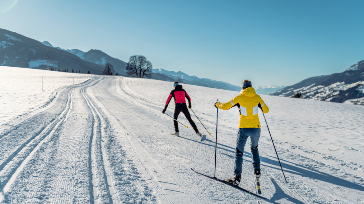 Zwei Personen beim Skilanglauf auf einer verschneiten Embach-Loipe in einer Berglandschaft unter strahlend blauem Himmel. (vergrößerte Ansicht)