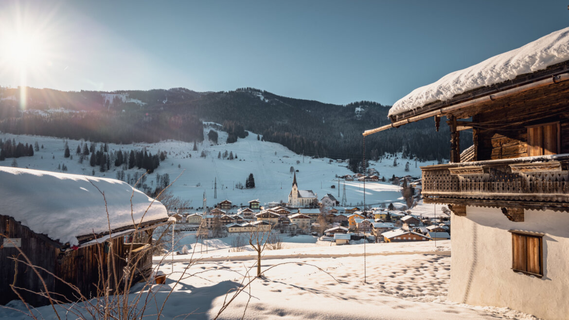 Verschneites Alpendorf Embach mit Holzhäusern, einer Kirche und sonnenbeschienenen Schneebergen im Hintergrund. (vergrößerte Ansicht)