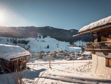 Verschneites Alpendorf Embach mit Holzhäusern, einer Kirche und sonnenbeschienenen Schneebergen im Hintergrund.