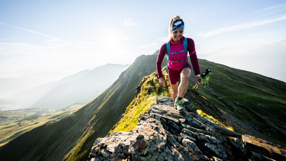 Frau beim Trailrunning entlang eines felsigen Bergrückens in Saalbach Hinterglemm, lächelnd und mit einer Wasserflasche in der Hand. (vergrößerte Ansicht)
