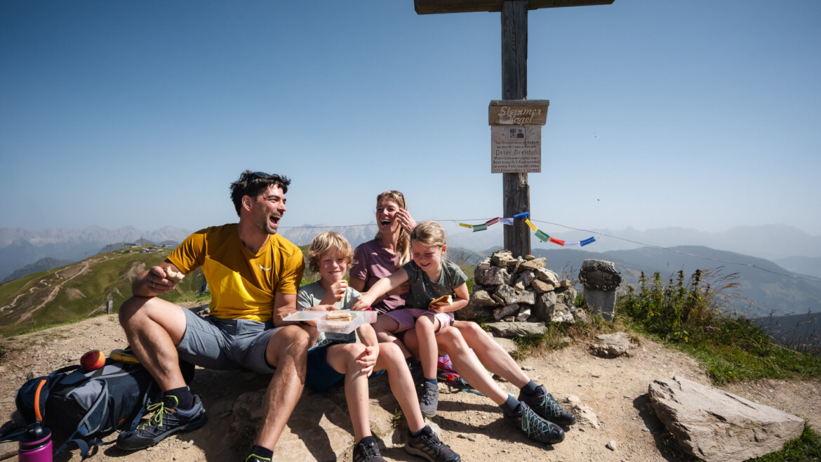 Eine vierköpfige Familie genießt eine Mahlzeit am Gipfelkreuz und bewundert die Aussicht auf die Berge von Saalbach Hinterglemm. (vergrößerte Ansicht)