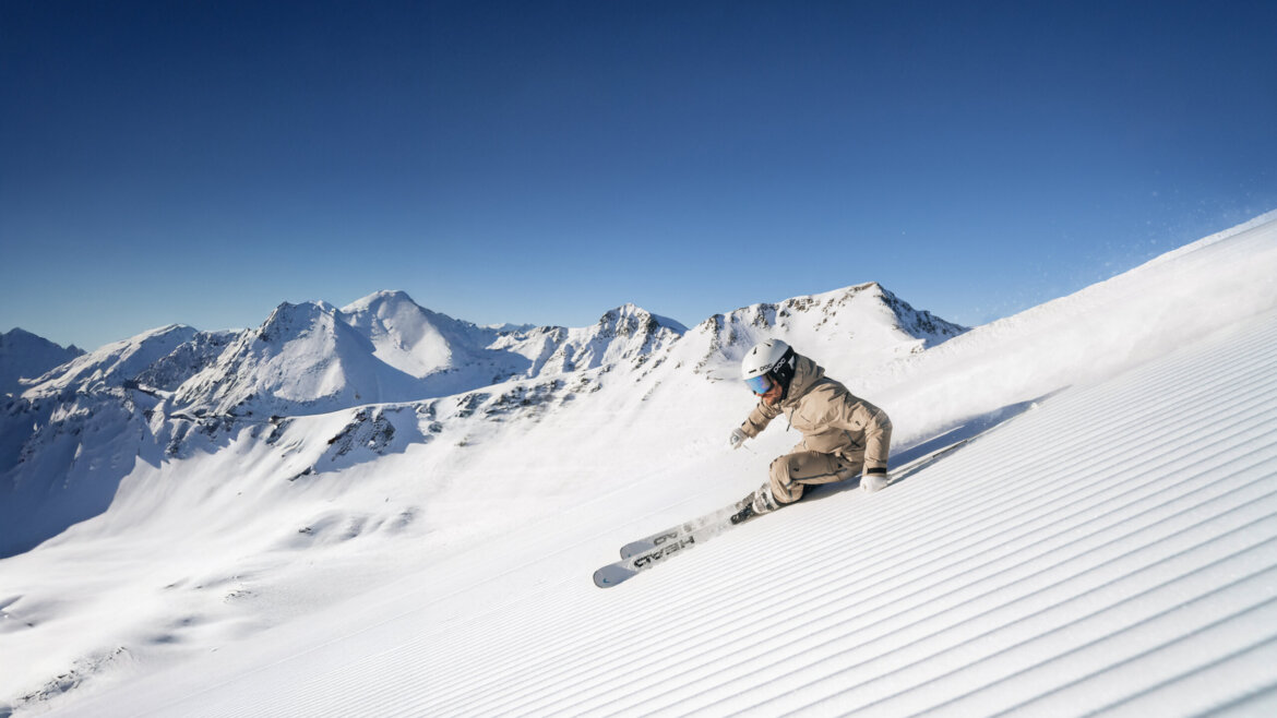 Ein Skifahrer fährt eine präparierte Piste in Saalbach Hinterglemm hinunter, umrahmt von schneebedeckten Gipfeln und einem strahlend blauen Himmel. (vergrößerte Ansicht)