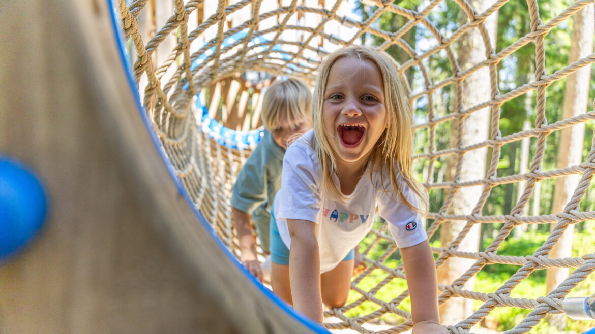 Zwei lachende Kinder krabbeln an einem sonnigen Tag durch einen Seiltunnel in Flachau, lachen und haben Spaß. (vergrößerte Ansicht)