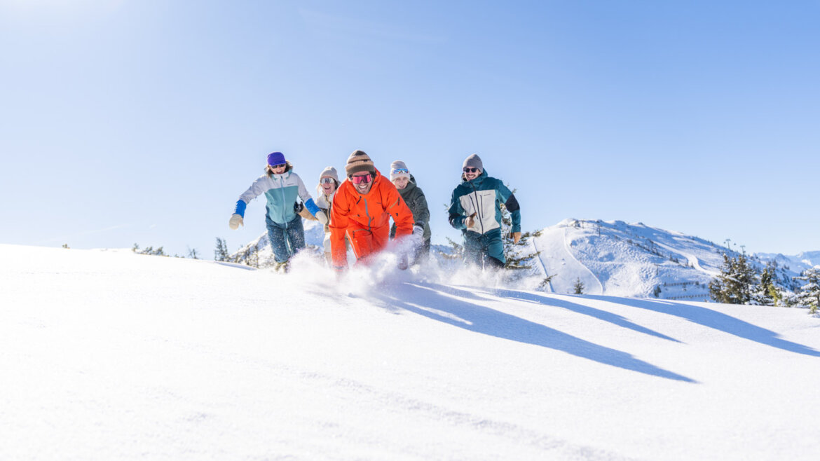 Fünf Personen in Winterkleidung laufen durch den Tiefschnee in Flachau, im Hintergrund leuchten verschneite Berge. (vergrößerte Ansicht)