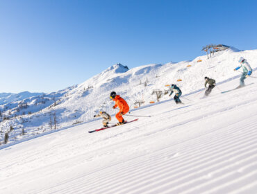 Vier Skifahrer fahren an einem sonnigen Tag eine verschneite Flachauer Piste hinunter, mit blauem Himmel und Skiliften im Hintergrund.