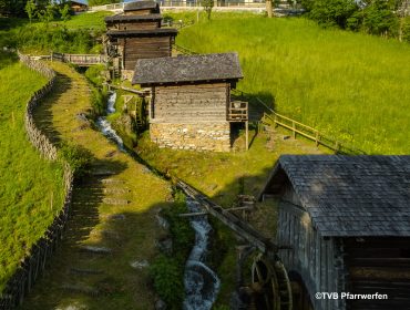 7 Mühlen Drei rustikale Holzmühlen mit Wasserrädern liegen am grasbewachsenen Hang von Pfarrwerfen, verbunden durch einen gewundenen Bach.