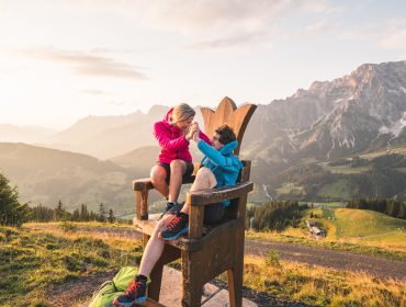A19_2300 Zwei Personen sitzen spielerisch auf einem riesigen Holzstuhl in Hochkönig, mit Blick auf Berge und ein grünes Tal bei Sonnenuntergang.