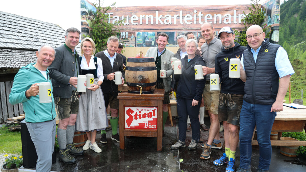 Zwölf Almsommer-Partner, die lächelnd mit Bierkrügen in der Hand auf einer Freiluftbühne stehen, vor der ein Bierfass und eine alpine Kulisse zu sehen sind. (vergrößerte Ansicht)