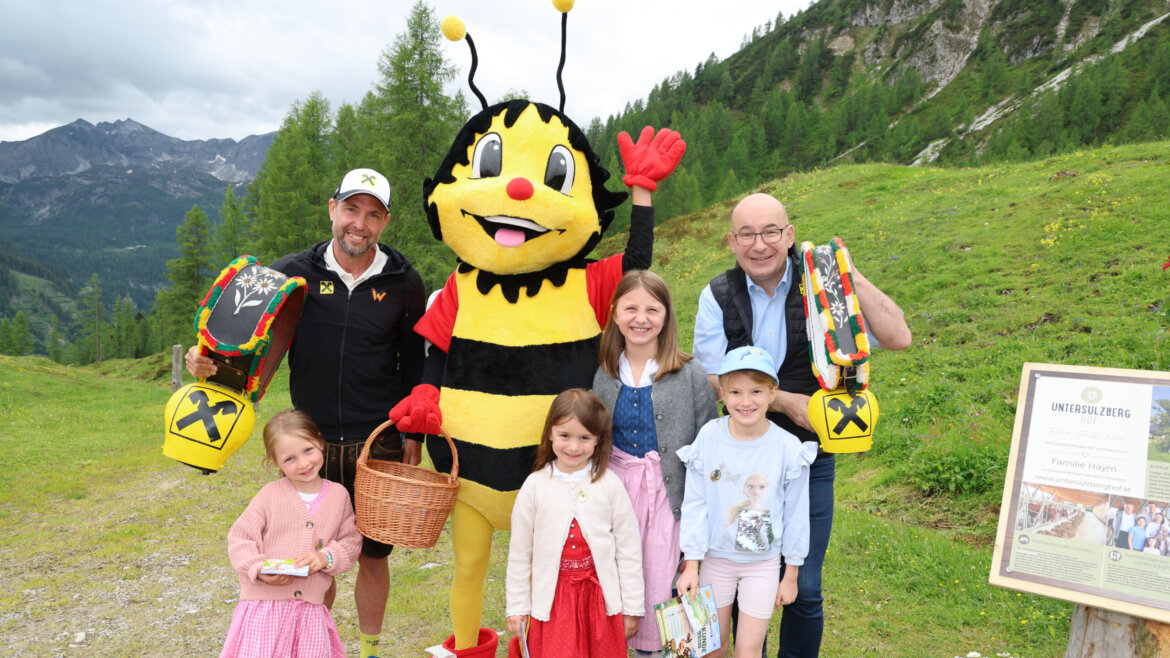 Eine Gruppe mit einer Biene als Maskottchen, Almsommer-Partner, lächelt draußen in den Bergen vor einem Schild und vor dem Hintergrund grüner Hügel. (vergrößerte Ansicht)
