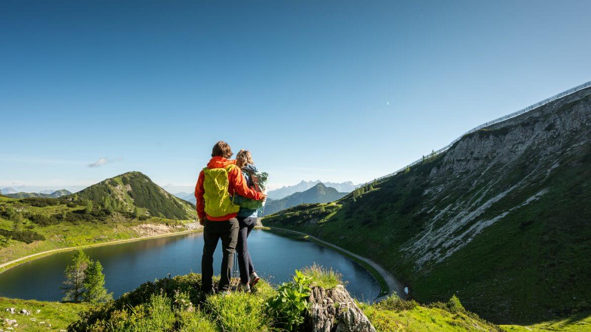 Zwei Wanderer mit bunten Rucksäcken stehen auf einem Hügel und blicken auf einen Bergsee, umgeben von grünen Bergen und blauem Himmel.