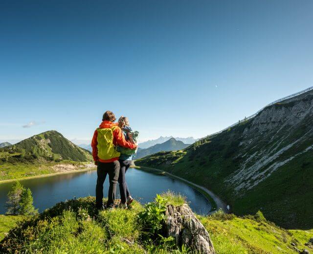Zwei Wanderer mit bunten Rucksäcken stehen auf einem Hügel und blicken auf einen Bergsee, umgeben von grünen Bergen und blauem Himmel.
