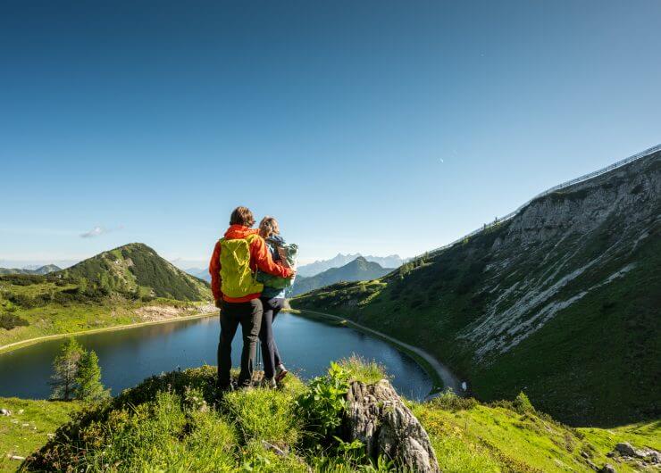 Zwei Wanderer mit bunten Rucksäcken stehen auf einem Hügel und blicken auf einen Bergsee, umgeben von grünen Bergen und blauem Himmel.