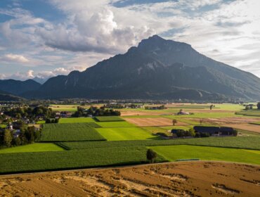 Anif © Gästeservice ANIF Luftaufnahme der Stadt Anif, die in einem Tal aus grünen Wiesen und goldenen Feldern unterhalb eines großen Berges liegt.