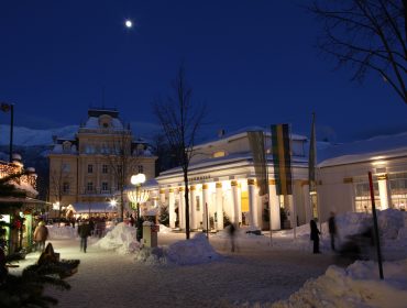 Der nächtliche Stadtplatz von Bad Ischl, mit schneebedeckten Straßen, beleuchteten Gebäuden und dem Mond am dunkelblauen Himmel.