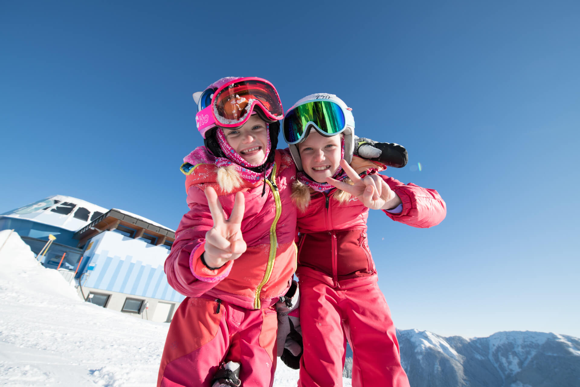 Zwei lächelnde Kinder in rosa Skikleidung machen in der Salzburger Sportwelt unter strahlend blauem Himmel Friedenszeichen.