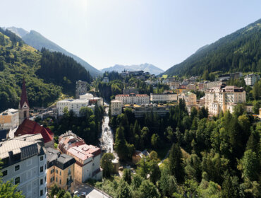 Ortszentrum Bad Gastein mit bunten Gebäuden, einer Kirche und dem Bad Gasteiner Wasserfall inmitten von grünen Hügeln und Wäldern.