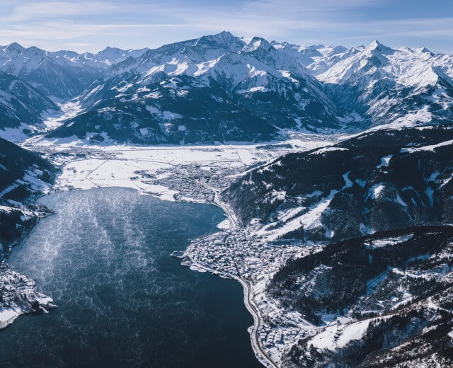 Luftaufnahme der verschneiten Berge und des zugefrorenen Sees von Zell am See mit einer kleinen Stadt entlang des Ufers.