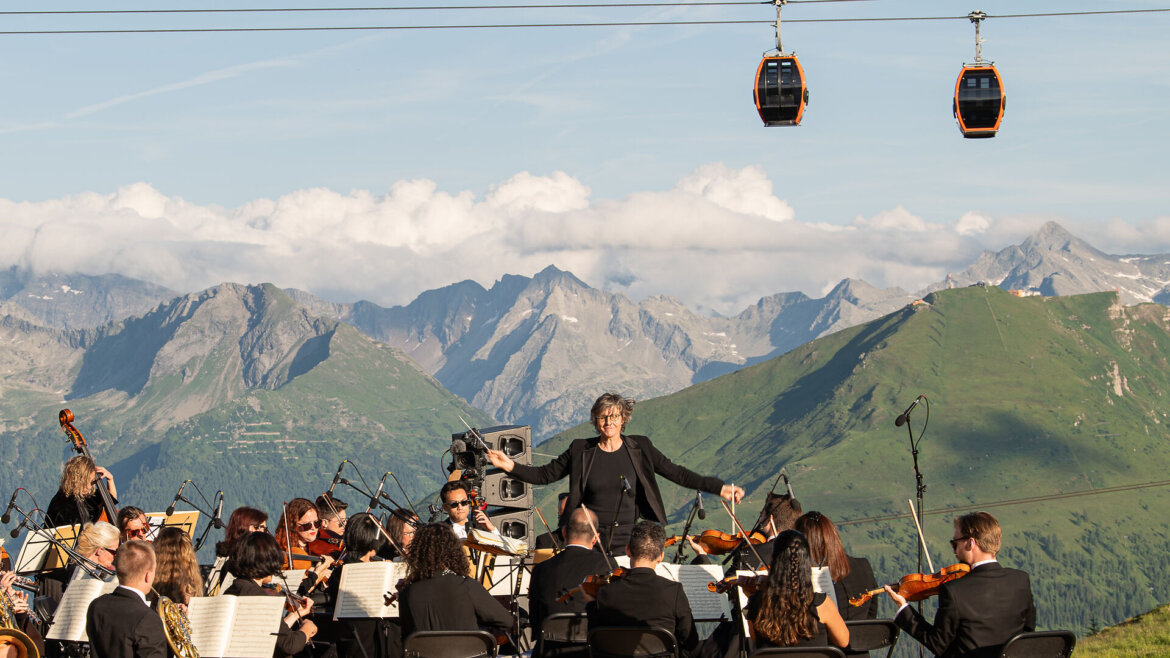 Ein Orchester spielt unter freiem Himmel in den Gasteiner Bergen, mit Seilbahnen und verschneiten Gipfeln im Hintergrund. (vergrößerte Ansicht)