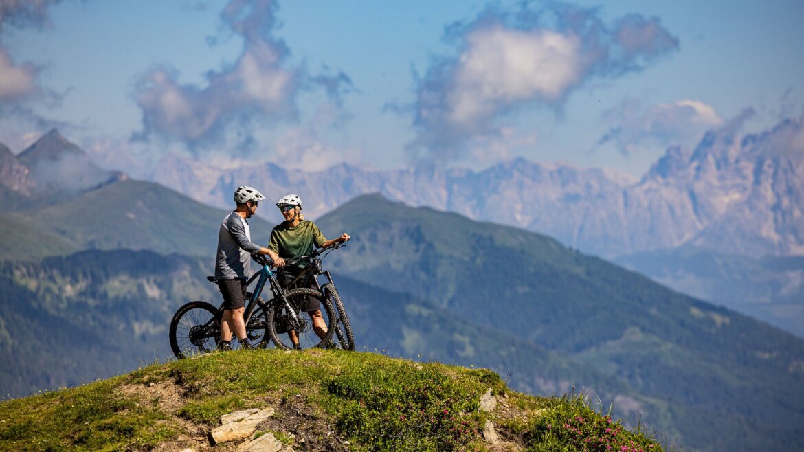 Zwei Radfahrer mit Mountainbikes stehen auf einer grasbewachsenen Gasteinertaler Bergkuppe, im Hintergrund sind Berge und Wolken zu sehen. (vergrößerte Ansicht)