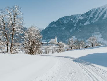Eine verschneite Landschaft mit Bäumen, einer Kirche und Bergen auf der Sonnenterrasse bei strahlend blauem Himmel.