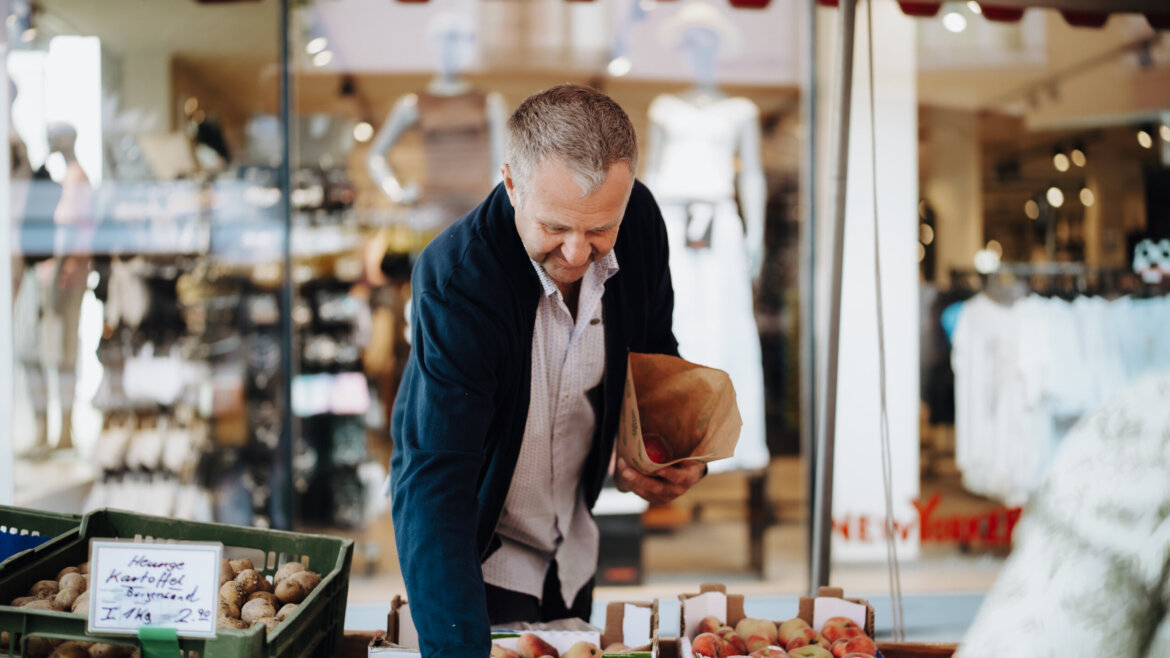 © Tourismusverband Bischofshofen | two minds Studio Ein Mann wählt auf einem Markt in Bischofshofen frische Produkte aus, hält eine Papiertüte in der Hand und lächelt. (vergrößerte Ansicht)