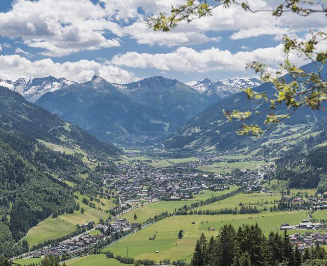 Das grüne Talstädtchen Bad Hofgastein ist umgeben von Bergen mit schneebedeckten Gipfeln unter einem teilweise bewölkten Himmel.