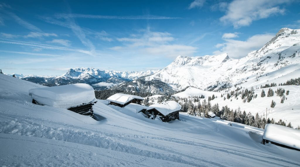 Verschneite Hütten an einem Berghang in Werfenweng, mit Kiefern und fernen schneebedeckten Gipfeln unter einem blauen Himmel.