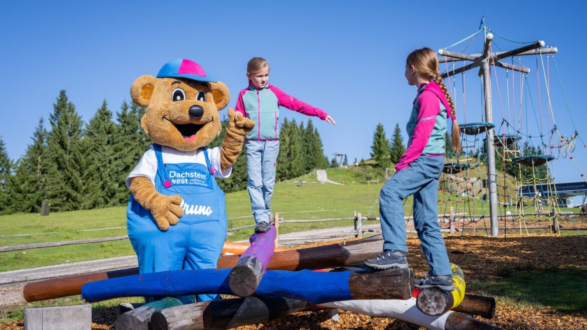 Ein Bärenmaskottchen und zwei Mädchen spielen auf einem bunten Balancierbalken auf dem Spielplatz in Dachstein West, der von Bäumen umgeben ist. (vergrößerte Ansicht)