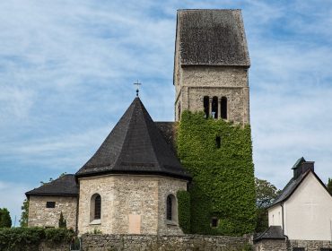 Eine Steinkirche mit einem hohen, efeubewachsenen Turm steht vor einem blauen Himmel mit leichten Wolken.