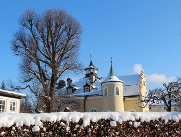 Elegantes gelbes Anif-Gebäude mit Türmen, verschneitem Dach, kahlen Bäumen und einer schneebedeckten Hecke unter blauem Himmel.
