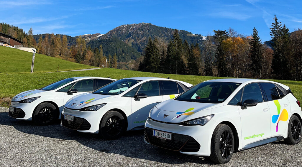 Drei weiße Elektroautos mit bunten Logos, die auf Schotter in Werfenweng geparkt sind, dahinter grüne Hügel und Berge.