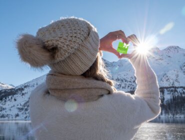 Eine Person in Winterkleidung rahmt mit ihren Händen die Sonne über einer verschneiten Berglandschaft eines österreichischen Biobauernhofs ein.