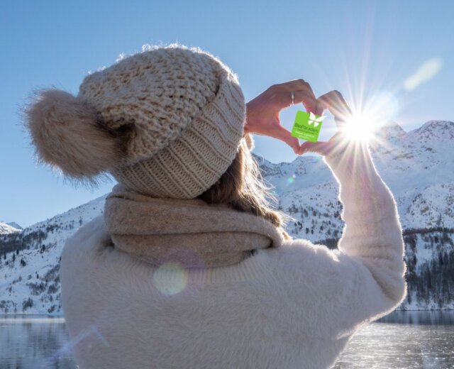 BioParadies Winter Titelbild Eine Person in Winterkleidung rahmt mit ihren Händen die Sonne über einer verschneiten Berglandschaft eines österreichischen Biobauernhofs ein.