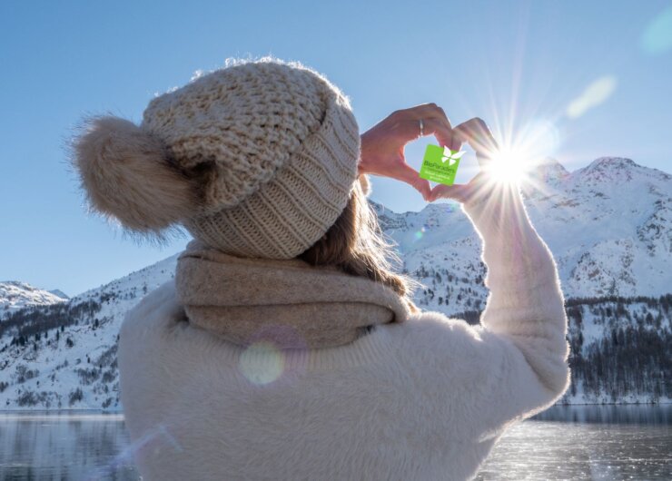 BioParadies Winter Titelbild Eine Person in Winterkleidung rahmt mit ihren Händen die Sonne über einer verschneiten Berglandschaft eines österreichischen Biobauernhofs ein.