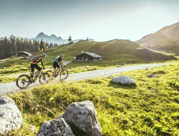 Zwei Radfahrer genießen das E-Biken auf einem Schotterweg durch malerische, sonnige Berge mit grünen Hügeln und Hütten.